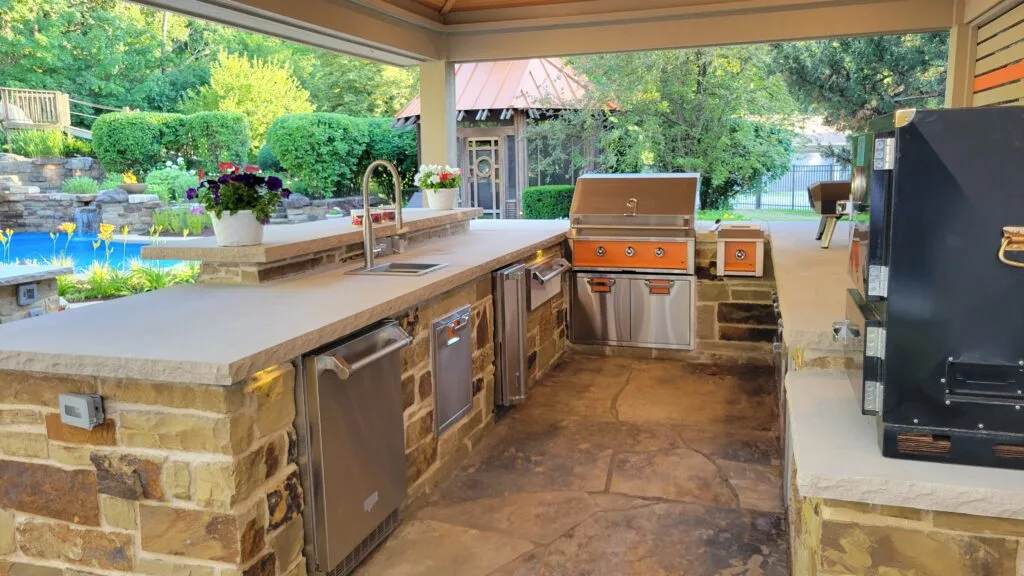 A stone outdoor kitchen with a built-in gas grill and smoker beside the pool